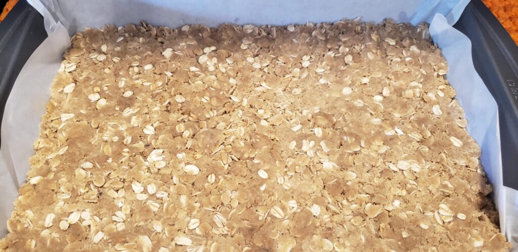 Unbaked oat crumble crust pressed into a parchment-lined pan for pumpkin pie bars